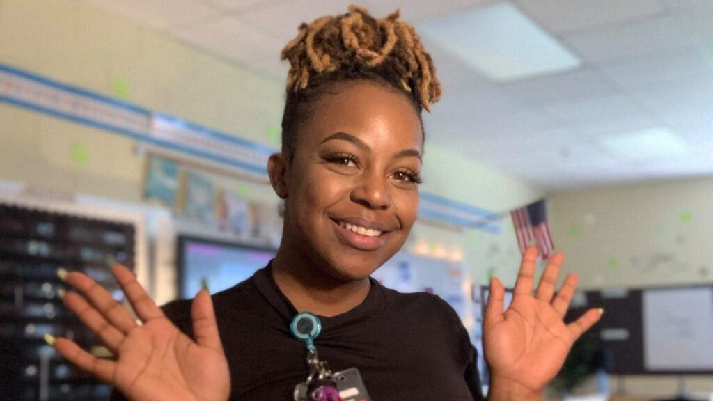 A woman standing in an empty classroom smiling with her hands fanned out.