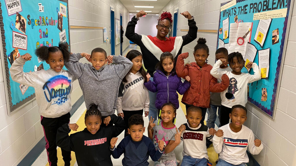 A group of second grade students with their female teacher in a school hallway.